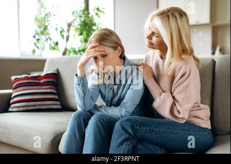 Mutter und Tochter zusammen im Wohnzimmer. Liebevolle reife Mutter sitzt auf der Couch und umarmt ihre junge Erwachsene Tochter, streichelt sie, sympathisiert, Familie Beziehung und Werte Konzept Stockfoto