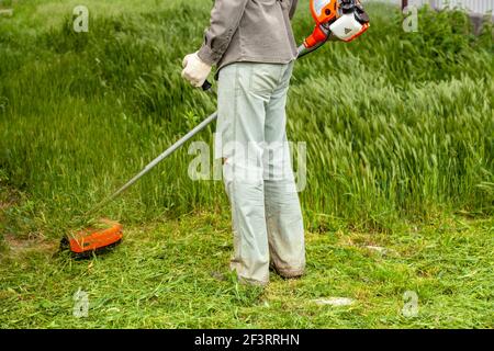 Ein Mann in Arbeitskleidung mäht hohes Gras mit einem Trimmer im Garten Stockfoto