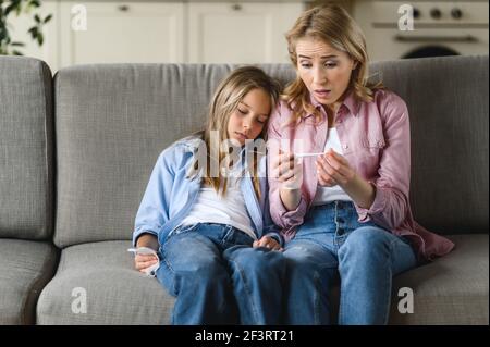 Mutter und Tochter zu Hause. Eine kaukasische, traurig besorgte Mutter sitzt auf einem Sofa, hält ein Thermometer und schaut ihn ängstlich an, während die ungesunde, kranke kleine Tochter neben ihr sitzt Stockfoto