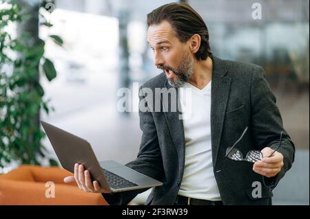 Stilvoller hübscher kaukasischer erwachsener Mann mit Laptop. Ein bärtiger, selbstbewusster Geschäftsmann in stilvoller Kleidung hält einen offenen Laptop in der Hand und schaut überrascht den Monitor an, indem er seine Brille auszieht Stockfoto