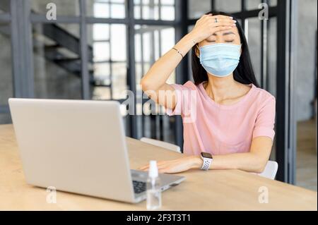 Junge asiatische Büroangestellte in Schutzmaske Kopfschmerzen, berühren Stirn und arbeiten auf Laptop, sitzen am Schreibtisch mit Desinfektionsmittel, soziale Distanzierung, zurück in das Büro in covid-19 Stockfoto