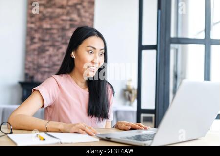 Junge asiatische Mischrennerin mit Ohrhörern, die am Schreibtisch sitzen, im Internet surfen und Nachrichten online lesen, auf den Laptop-Bildschirm schauen, kann ihren Augen nicht glauben und fühlt sich überrascht Stockfoto