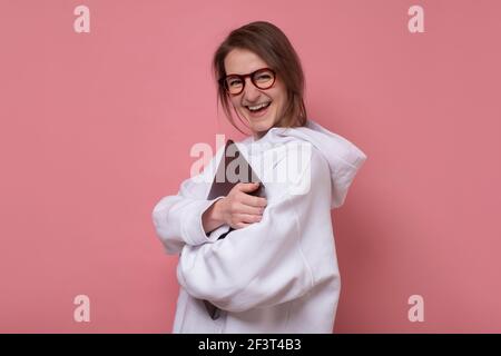 Lächelnde junge Frau in einer Brille, die mit einem Laptop gegen Rosa steht Hintergrund Stockfoto