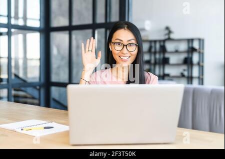 Intelligente asiatische Managerin in Brille sitzt am Schreibtisch im modernen Büro, winkt am Laptop, trägt Kopfhörer, Mixed-Race-Frau spricht mit Kunden online, Projekt mit Kunden zu diskutieren Stockfoto