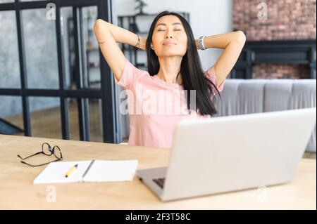 Erfolgreiche junge asiatische Geschäftsfrau entfernt Gläser sitzen am Schreibtisch mit Laptop, Gefühl aufgeregt für die Fertigstellung des Projekts, legte die Hände hinter den Kopf, Stretching, bewundern eigene Arbeit, getan arbeiten Stockfoto