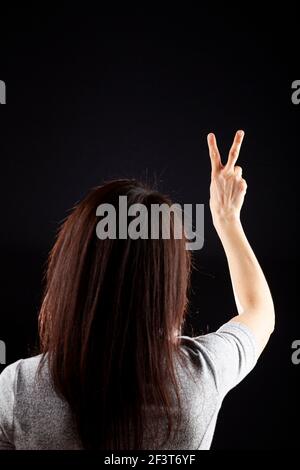 Hinter dem Kopf Blick auf eine junge kaukasische Frau mit langen braunen Haaren trägt Kurzarm-Shirt vor dunklem Hintergrund. Sie zeigt zwei Finger mit ihm Stockfoto