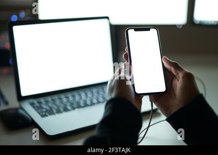 Verwenden von Minianwendungen. Nahaufnahme des weißen Bildschirms von Smarthone. Mann, der im dunklen Raum vor vielen Computermonitoren sitzt, während er spät alleine arbeitet Stockfoto