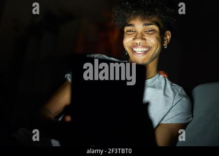 Porträt von fröhlichen jungen Kerl lächeln, während mit Tablet-pc in der Nacht, sitzen in dunklen Raum zu Hause Stockfoto