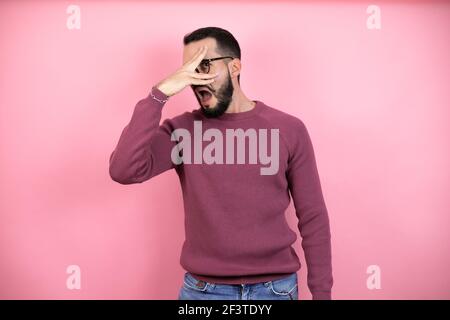 Schöner Mann trägt Brille und Freizeitkleidung über rosa Hintergrund in Schock schauend Gesicht und Augen mit der Hand, Blick durch die Finger mit e Stockfoto