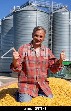 Lächelnder Maisfarmer beim Blick auf Kamera gegen Stahlkornsilos. Traktor mit Fresly geernteten Maiskörnern neben Maistrocknersilos beladen. Stockfoto