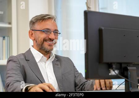 Erfolgreiche Reife Geschäftsmann sitzen am Office Desk. Lächelnder, eleganter Mann, der in seinem Büro Computermonitor anschaut. Stockfoto