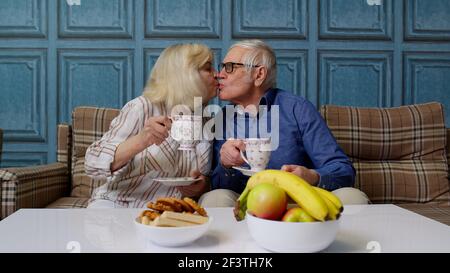 Seniorenpaar hält Tee trinken reden, lachen, lächeln, küssen, sitzen auf der Couch zu Hause Stockfoto