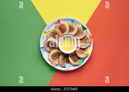 Teller mit kleinen runden Pfannkuchen mit bunten Party Topper Flaggen auf Split dreifarbig geschichteten Papier Hintergrund. Draufsicht, kräftige, lebendige Farben, Orange Stockfoto