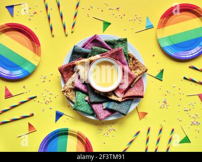 Rainbow Party, Tischaufbau. Lebendige Crepes auf Teller mit Honig. Partytisch, gelbe Papiertischdecke, Trinkhalme, Flaggen, Regenbogen-Pappteller Stockfoto