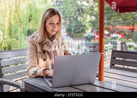 Junge kaukasische Geschäftsfrau mit blonden Haaren, die in einem Café im Freien auf einem Laptop arbeiten. College-Student mit Technologie , Online-Bildung, freiberuflich Stockfoto