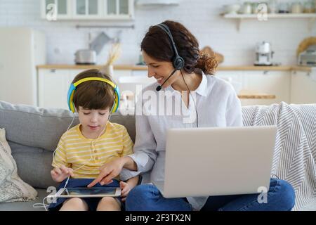 Glückliche Mutter mit Kind sitzt auf dem Sofa zu Hause während Lockdown Arbeit auf Laptop Kind spielen im Tablet Stockfoto