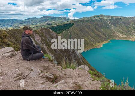 Männlicher Tourist mittleren Alters mit Blick auf die türkisfarbene Quilotoa vulkanische Lagune vom höchsten Gipfel entlang der Rundwanderung. Stockfoto
