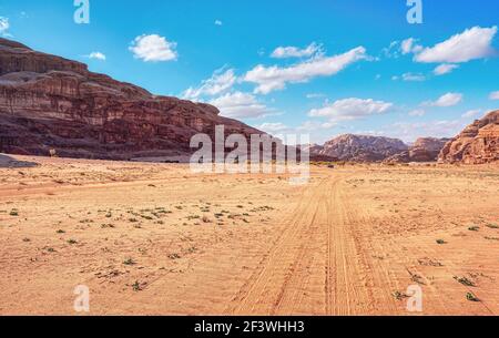 Felsige Massive auf roter Sandwüste, heller wolkiger Himmel im Hintergrund, kleines 4WD Fahrzeug und Kamel in der Ferne - typische Landschaft im Wadi Rum, Jordanien Stockfoto