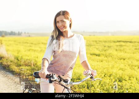 Junge Frau, die neben ihrem Fahrrad über staubige Landstraße steht, starke nachmittägliche Sonnenbeleuchtung im Hintergrund leuchtet auf gelbem Blumenfeld Stockfoto