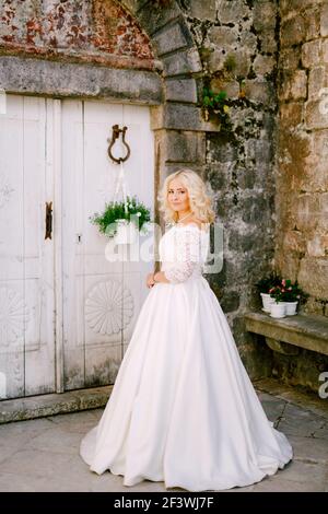 Eine Braut steht vor einem alten Backsteingebäude In der Nähe einer weißen Holztür mit Blumentöpfen in Perast Stockfoto