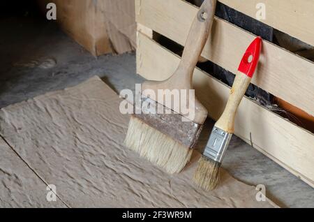 Alte Pinsel stehen neben der Holzkiste. Zwei Bürsten verschiedener Größen stehen auf dem Boden, der mit schmutziger Pappe bedeckt ist. Seitenansicht aus einem Winkel. Se Stockfoto