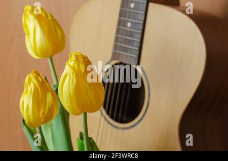 Blumenstrauß und eine akustische Gitarre auf braunem Hintergrund. Drei gelbe Tulpen aufrecht. Verblassende Popularität, vergessener Ruhm Konzept. Stockfoto