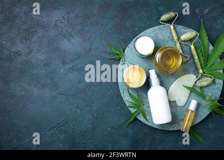Natürliche Haut und Selbstpflegekonzept. Flach liegend. Hanfblätter, Öl, Kosmetikprodukte, Gesichtscreme, Körperbutter, Gesichtswalze und Gua Sha Massagegerät im Dunkeln Stockfoto