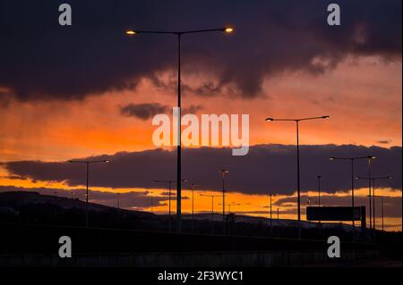 Spektakulärer roter Sonnenuntergang mit dunklen Regenwolken über der Brücke mit Silhouetten von Menschen und Fahrzeugen, hohe Straßenlaternen entlang des Highway M50 Dublin Stockfoto