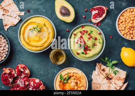 Schalen mit gelbem Hummus und grünem Hummus, Tahini, Olivenöl, Sesamsamen, Pita, Rohe Kichererbsen, Avocado, Granatapfel auf dunklem Grund. Mitte Stockfoto