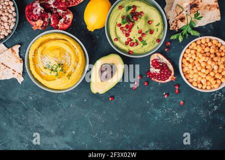 Schalen mit gelbem Hummus und grünem Hummus, Tahini, Olivenöl, Sesamsamen, Pita, Rohe Kichererbsen, Avocado, Granatapfel auf dunklem Grund. Mitte Stockfoto
