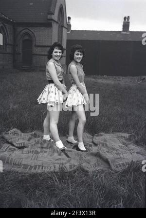 1958, historische, frühlingshafte und zwei Mädchen im Teenageralter, die die junge weibliche Mode des Tages tragen, kurze Polka-Punkt-Röcke, weiße Socken und kurzärmelige Oberteile, die auf einem Teppich für ihr Foto auf dem Gelände einer Kirche stehen, bevor sie an der Parade und dem Maifest teilnehmen, Leeds, England, VEREINIGTES KÖNIGREICH. Stockfoto