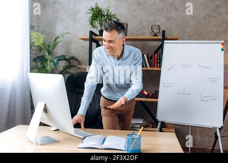 Selbstbewusster kaukasischer Geschäftsmann im Büro. Der Erwachsene Business Leader in formeller Kleidung führt Online-Briefing oder -Präsentation für Kollegen durch, die Computer verwenden, zeigt die Finanzstrategie auf dem Flipchart Stockfoto