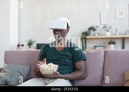 Afrikanischer Mann mit vr Gläser sitzen auf Sofa essen Popcorn Und Filme online ansehen Stockfoto