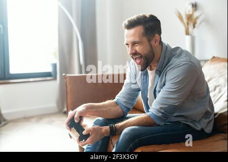 Porträt des jungen bärtigen Hipster Kerl sitzt auf der Couch im Wohnzimmer, halten Joystick, schreien, und gewinnen ein Videospiel-Match, männlich süchtig Gamer mit einem offenen Mund ist im Begriff, erfolgreich zu sein Stockfoto