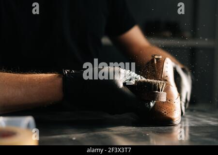 Nahaufnahme Hände von Schuhmacher tragen schwarze Handschuhe streicht Staub von der Schuhbürste in dunklen Handwerk Schuhgeschäft. Stockfoto