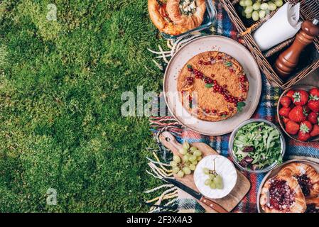 Picknick-Strohkorb mit gesunden Lebensmitteln, Zubehör. Sommerpicknick mit Kuchen, Obst, Käse, Wein und Snacks auf karierten grünen Hintergrund. Draufsicht Stockfoto