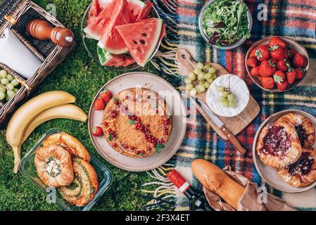 Picknick-Strohkorb mit gesunden Lebensmitteln, Zubehör. Sommerpicknick mit Kuchen, Obst, Käse, Wein und Snacks auf karierten grünen Hintergrund. Draufsicht Stockfoto