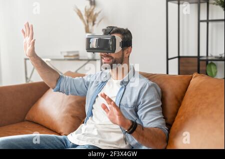 Glücklicher junger bärtiger Hipster, der auf dem Sofa im Wohnzimmer sitzt, zu Hause die Virtual-Reality-Technologie erlebt, in modernen VR-Brillen Spiele spielt und mit den Händen im Cyberspace drückt Stockfoto
