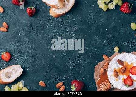Französischer Camembert-Käse, serviert auf Trauben, Honig, Mandeln, Erdbeeren und Baguette auf dunklem Grund. Draufsicht. Speicherplatz kopieren. Romantische Dattsnacht Stockfoto