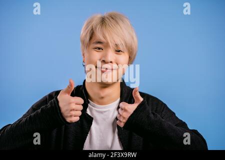 Schöner koreanischer Mann zeigt Daumen nach oben, wie Geste. Glücklich asiatische Kerl auf blauem Hintergrund. Gewinner. Erfolg. Positive männliche Modell lächelt zur Kamera. Körper Stockfoto