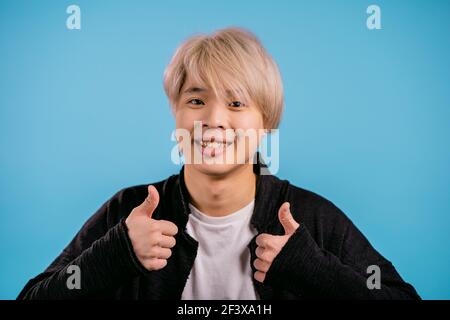 Schöner koreanischer Mann zeigt Daumen nach oben, wie Geste. Glücklich asiatische Kerl auf blauem Hintergrund. Gewinner. Erfolg. Positive männliche Modell lächelt zur Kamera. Körper Stockfoto