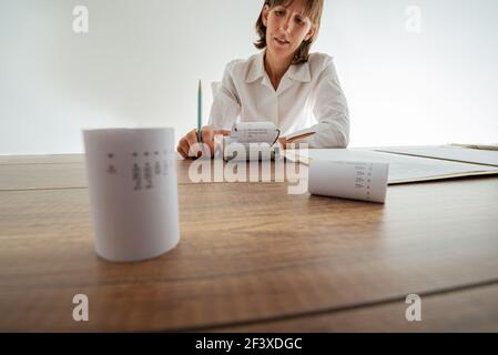 Junge Buchhalterin, die eine zusätzliche Maschine benutzt, die an ihrem Schreibtisch voll mit Quittungen und Dokumenten arbeitet. Stockfoto