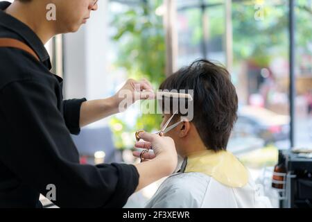 Barber trimmt die Haare eines jungen Mannes mit einer Schere In seinem Friseurladen Stockfoto