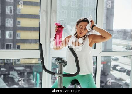 Eine junge Frau mit gemischtem Rennen wischt ihr Gesicht mit einem Handtuch, erschöpft von einem schweren Cardio-Training. Zeit zum Ausruhen nach dem Ergometer Stockfoto