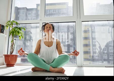 Junge afroamerikanische Frau, die in Lotuspose mit Händen in Ahamkara Mudra sitzt. Frau meditiert zu Hause an einem schönen sonnigen Tag Stockfoto