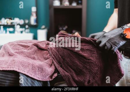 Professioneller Barbier wendet heißes Handtuch auf das Gesicht des männlichen Kunden, um Bart vor der traditionellen Rasurrasur, Nahaufnahme zu entspannen Stockfoto