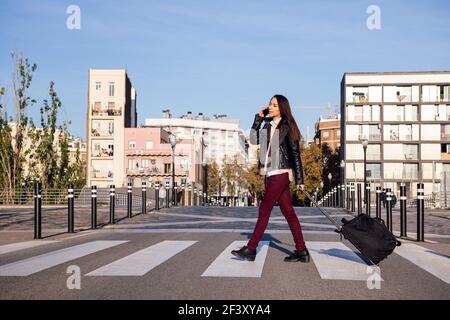 Junge Frau zieht Koffer telefonieren beim Überqueren der Straße an einem Fußgängerüberweg, Konzept von Reisen und urbanem Lebensstil, Copyspace fo Stockfoto