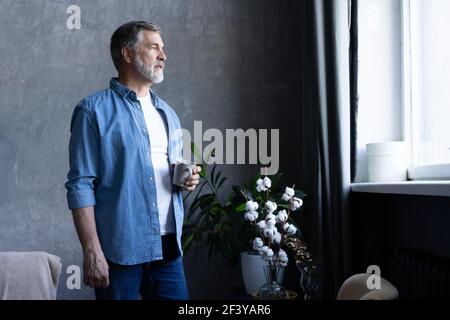 Ein gutaussehender älterer Mann hält eine Tasse, schaut weg und lächelt, während er in der Wohnung steht. Stockfoto
