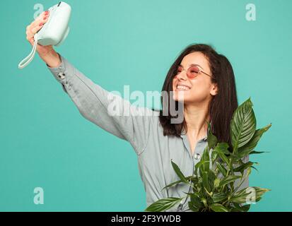 Foto von attraktiven Dame in Sonnenbrille hält Kamera, Pflanzen und ein Selfie. Trägt graues Hemd, isoliert türkisfarbenen Hintergrund Stockfoto