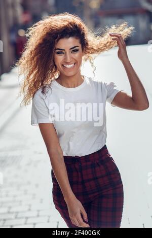 Lächelnden jungen arabischen Frau mit schwarzen Locken Frisur. Arabische Mädchen in Freizeitkleidung in der Straße. Gerne weibliche tragen weiße T-Shirt und Hose. Stockfoto